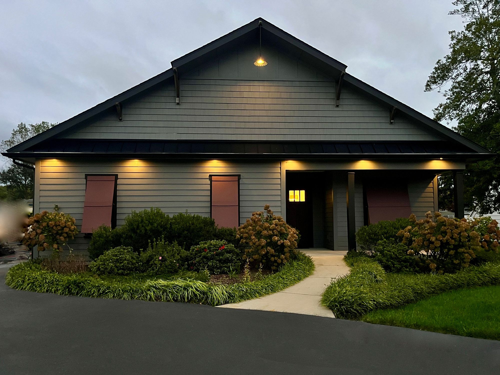 The Retreat on Savannah at dusk: a gray craftsman-style single-story home with rust-red shutters, lit soffits and a curving front walk through hydrangeas
